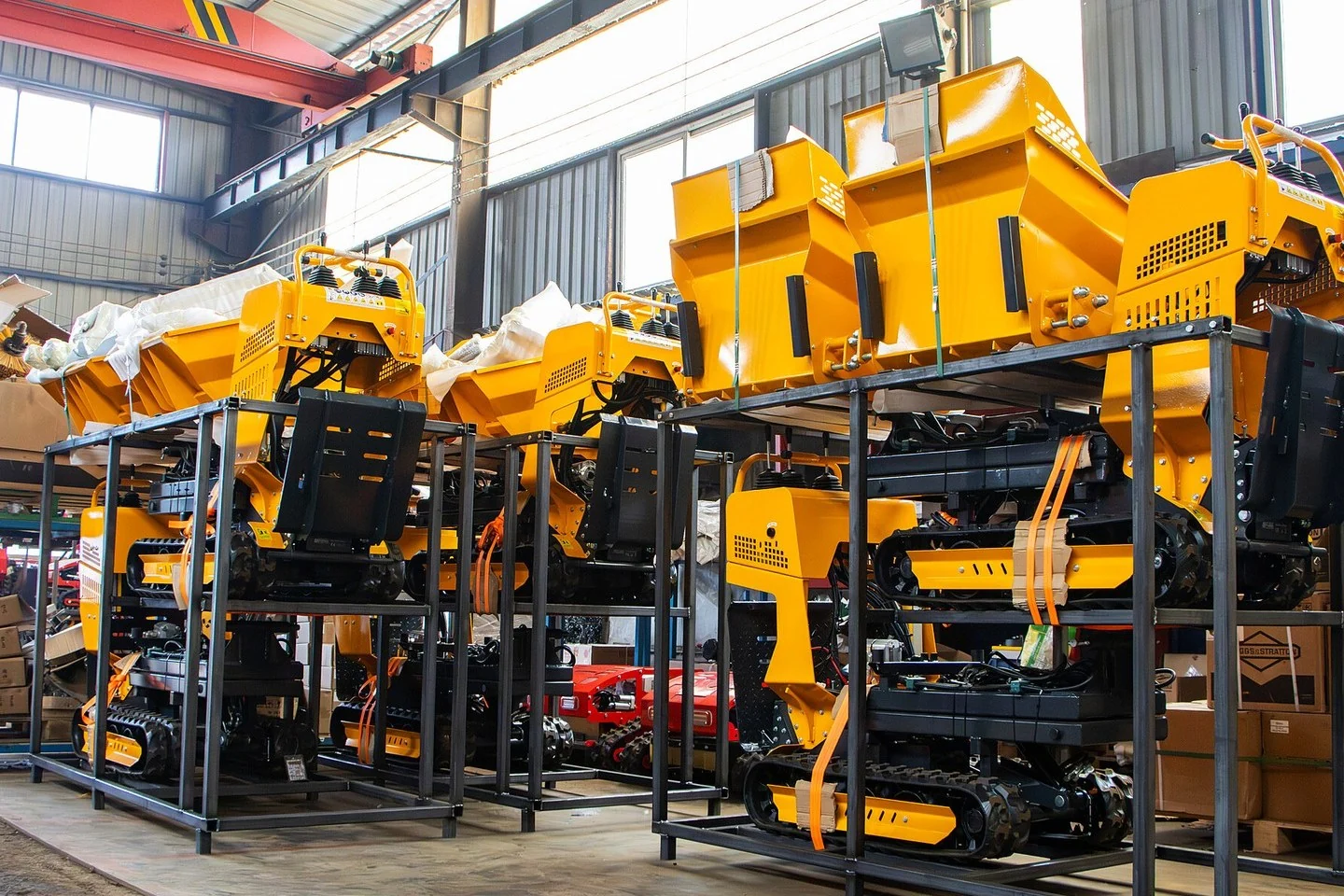 Finished yellow tracked mini dumpers strapped on black steel loading racks, two tiers high, in a factory hall