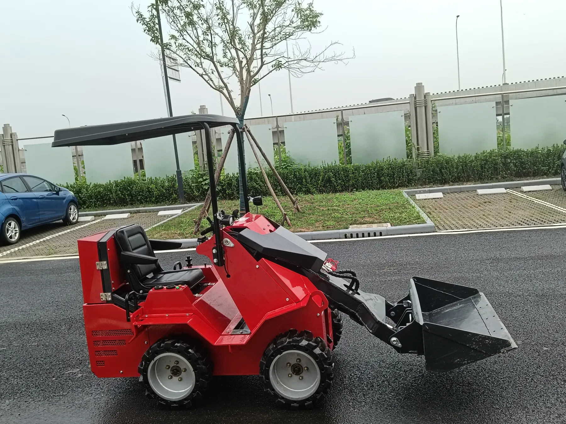 Red BL-S390-C wheeled seated mini skid steer from the side, with a black seat, a sun canopy on a frame and a black bucket lowered, parked on wet asphalt
