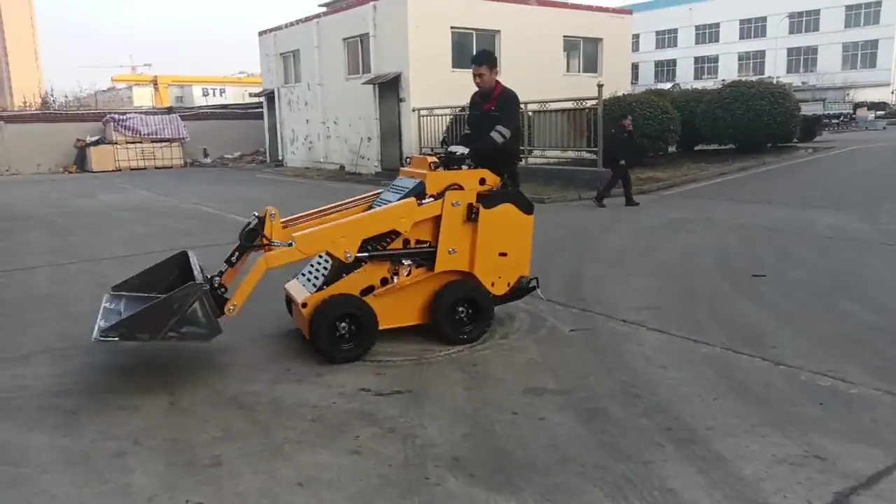 Operator standing on a yellow wheeled stand-on mini skid steer turning on a concrete yard with its bucket raised, factory buildings behind