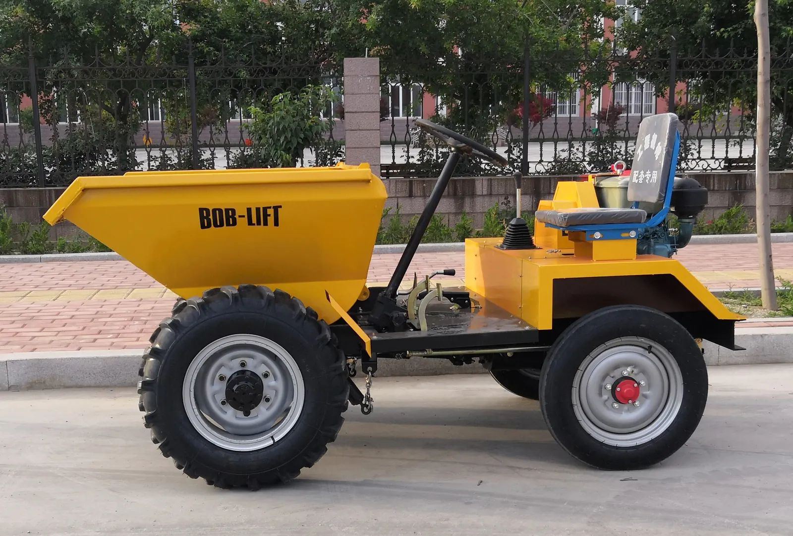 Yellow FC10 wheeled mini dumper in side view, with a front tipping hopper marked BOB-LIFT, a steering wheel and an open seat behind it, parked on a paved road