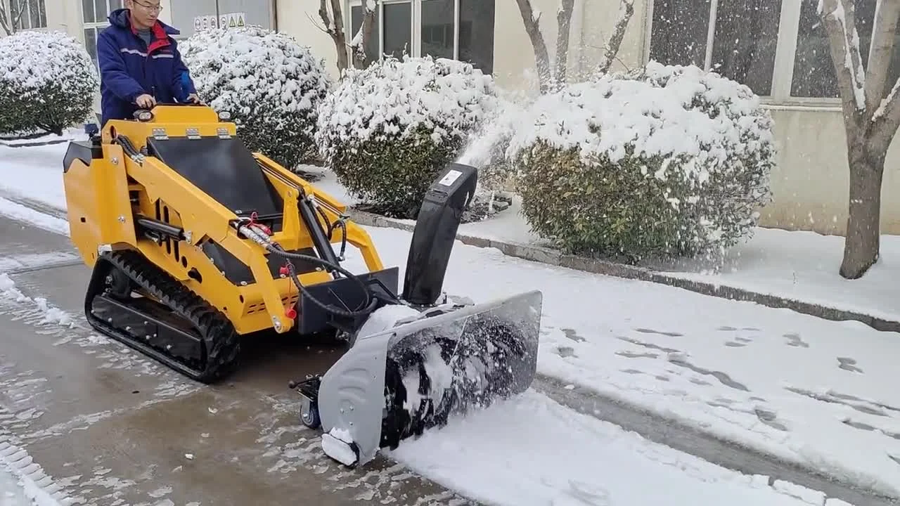 Yellow BL-T750-H tracked stand-on mini skid steer with a snow blower attachment throwing snow off a cleared road beside snow-covered hedges